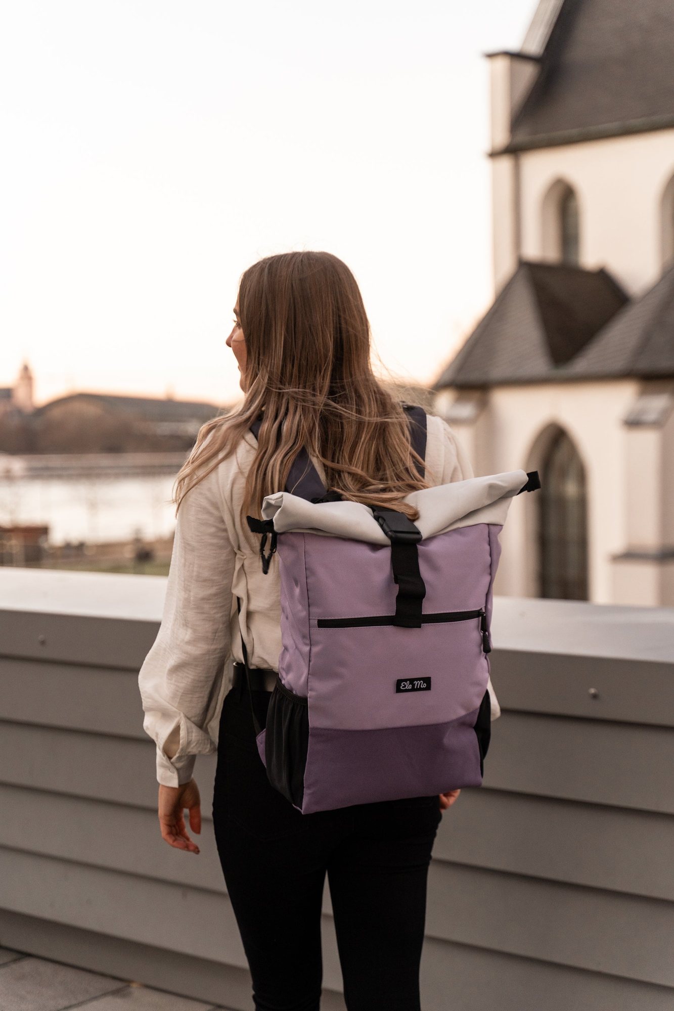 Ela Mo – Rückenansicht einer Person mit lavendelfarbenem Ela-Mo-Rolltop-Rucksack vor einer Kirche (offizielles Produktfoto)